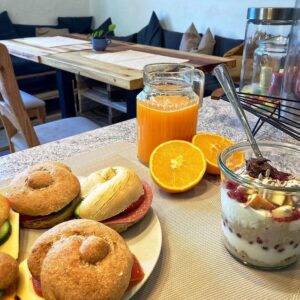 Ein Tisch gedeckt mit belegten Brötchen, Müsli und frischem Orangensaft. Im Hintergrund eine Sitzecke im Cafe.