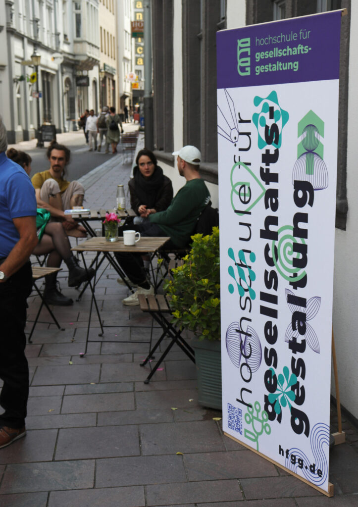 Ein Foto eines Aufstellers der Hochschule für Gesellschaftsgestatung: Im HIntergrund sitzen an einem Tisch, auf einer Straße vier junge Menschen.