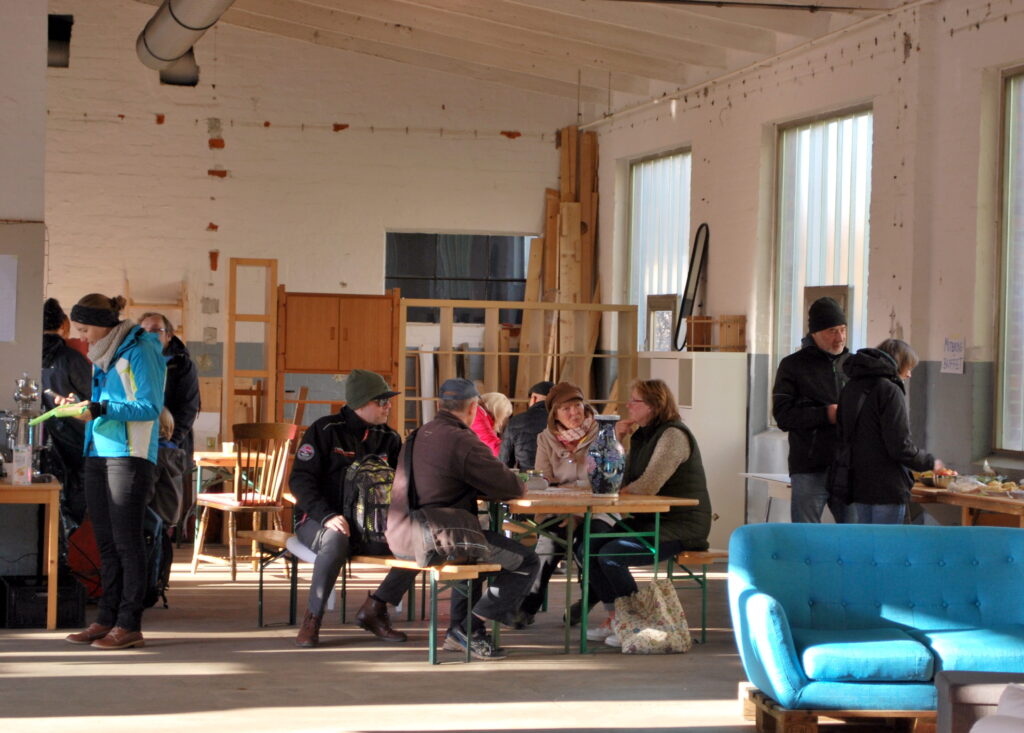 Ein Foto aus der Halle des Commons Zentrums. Einige Menschen sitzen an einem Tisch und unterhalten sich, andere bedienen sich am Buffet