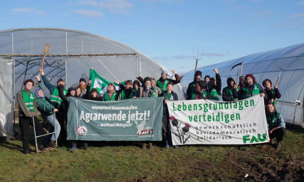 Vor Folientunnel stehen Aktivist:innen in grünen FAU-Westen; einige erheben die Hände zu kämpferischen Fäusten. Auf den Transparenten steht u.a. "Agrarwende jetzt!" und "Lebensgrundlagen verteidigen".