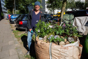 Eine besondere Form des Urban Gardening: Ein alter, großer Sandsack auf einem Fußweg, der als Hochbeet umfunktioniert wurde. Dahinter steht eine Frau mit Gartenschere.