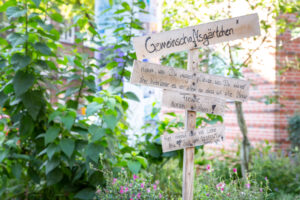 In einem schönem Garten, vor einem Backsteinhaus, steht ein Schíld mit der Aufschrift: Gemeinschaftsgärtchen. Urban Gardening.