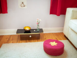 Zu sehen ist ein rotes Meditationskissen vor einem kleinem Schränkchen, mit Blumen und einem Teelicht, in einer goldenen Kokusnuss-Schale.